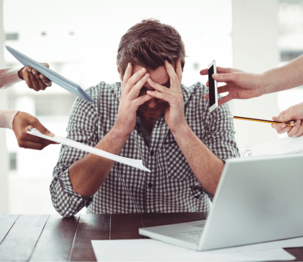 A stressed worker holds his head in his hands while multiple colleages try to give him more work.